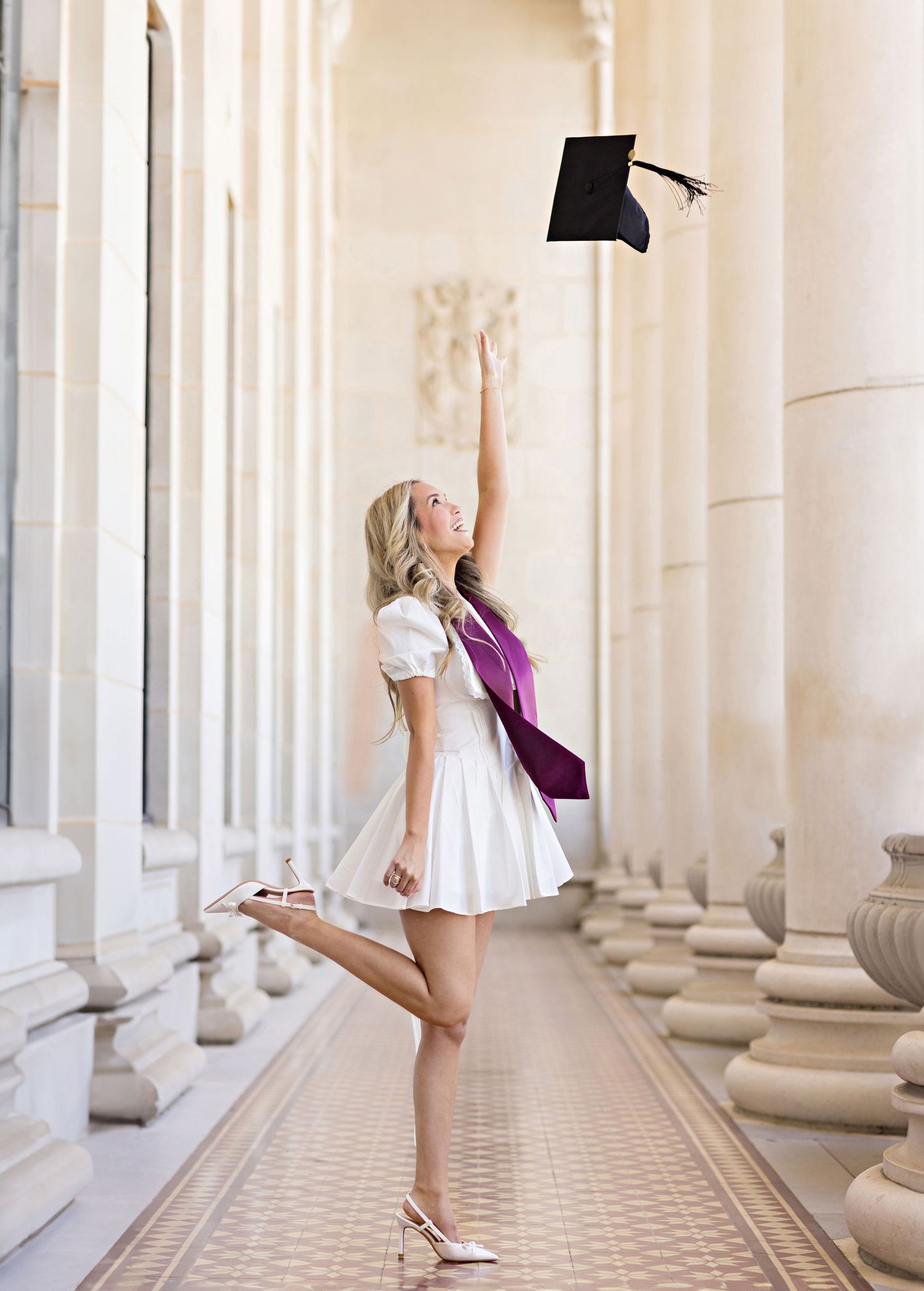 A graduate in a white dress celebrates by tossing their cap in an elegant colonnade hallway.