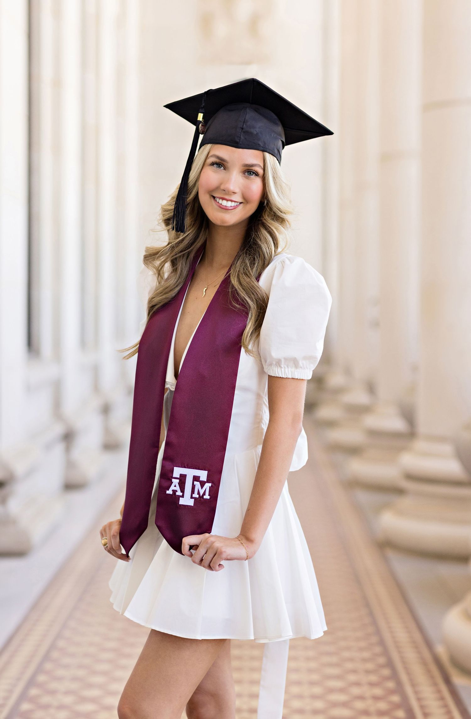 Graduate wearing white dress and maroon Texas A&M stole poses in front of columns during graduation photo session.