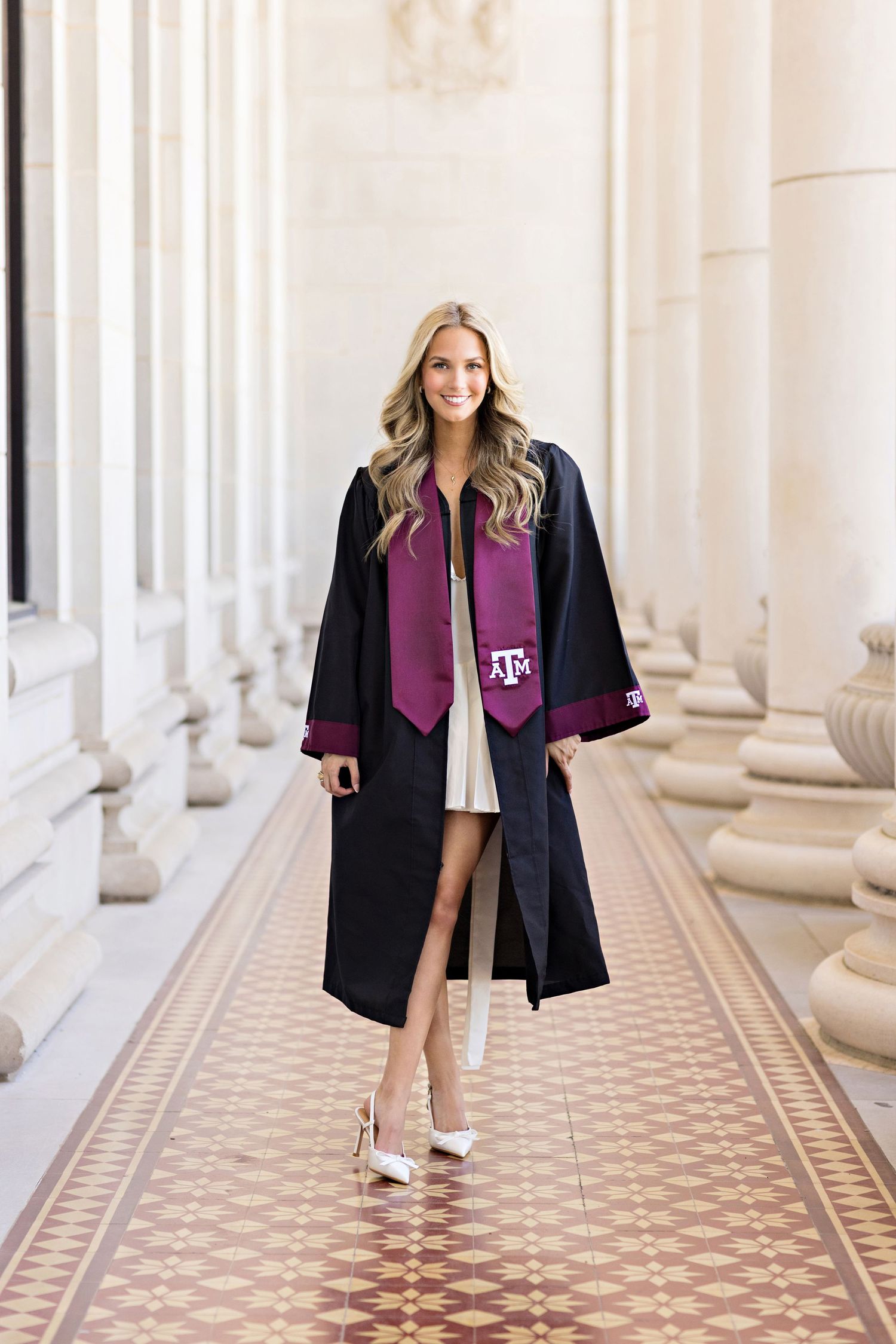 Graduate wearing traditional black graduation gown and stole walking along ornate tiled corridor.