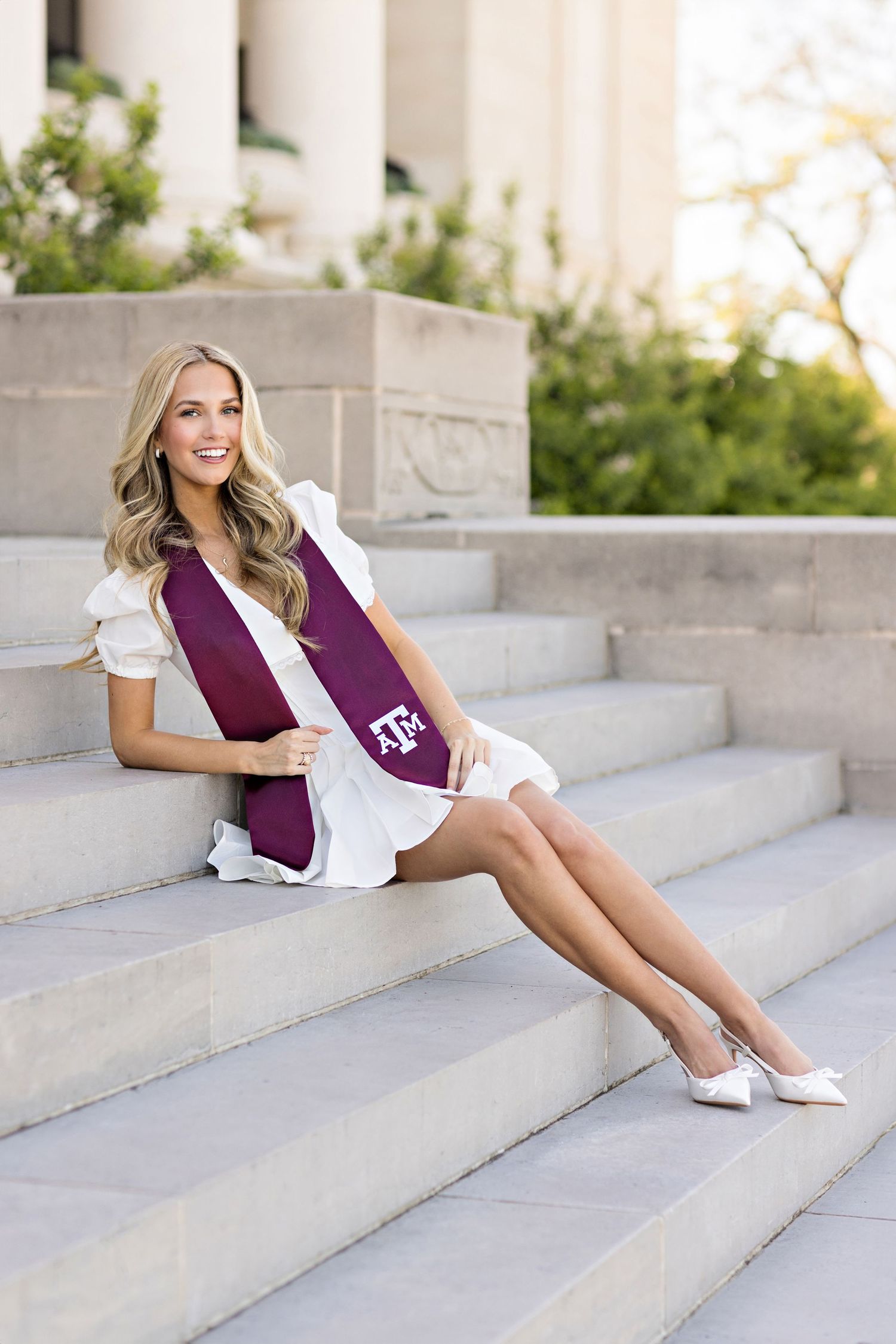 A graduate wearing a white dress sits on stone steps with maroon Texas A&M sash for senior portraits.