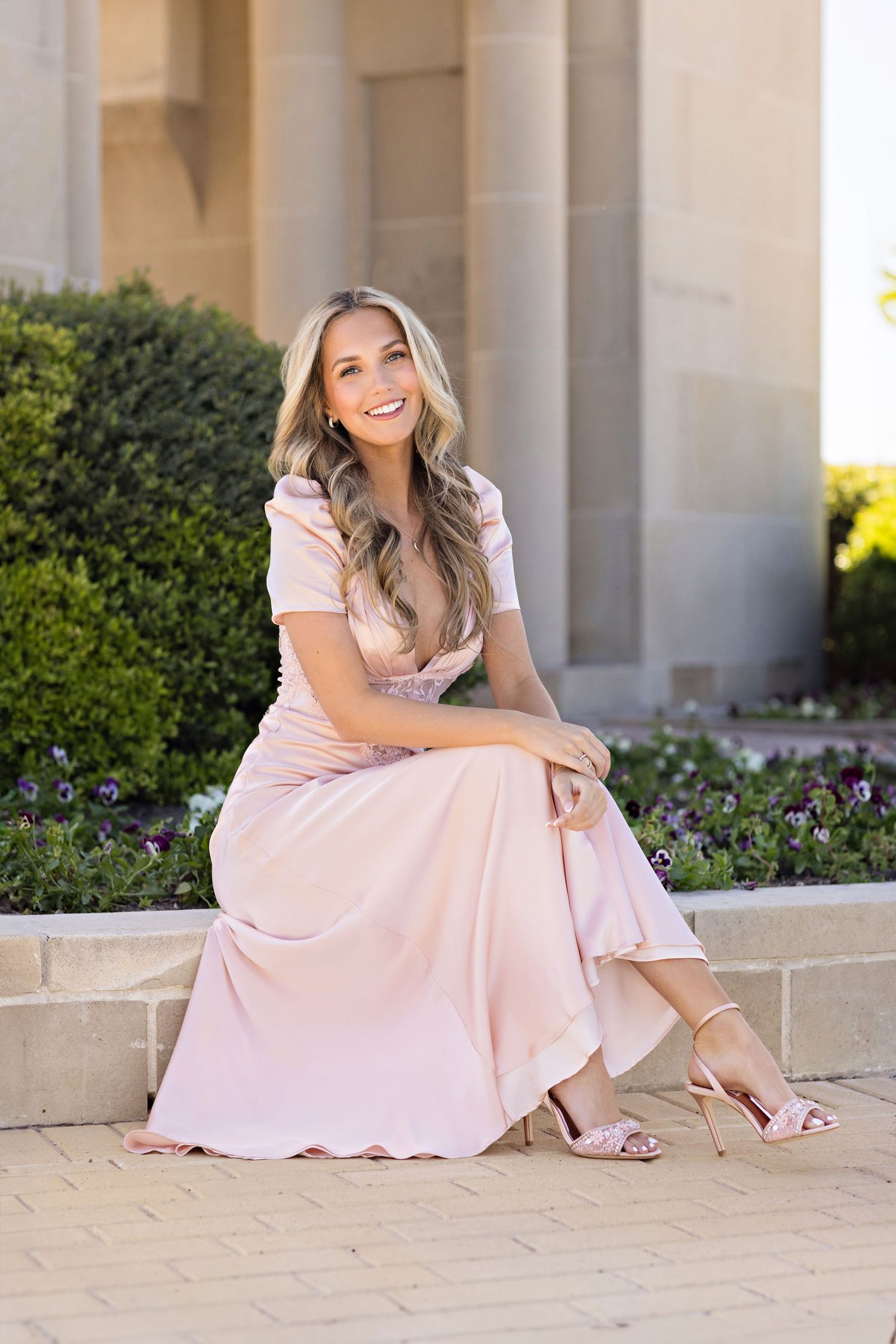 Woman in light pink maxi dress sitting elegantly on stone steps among garden flowers and columns.