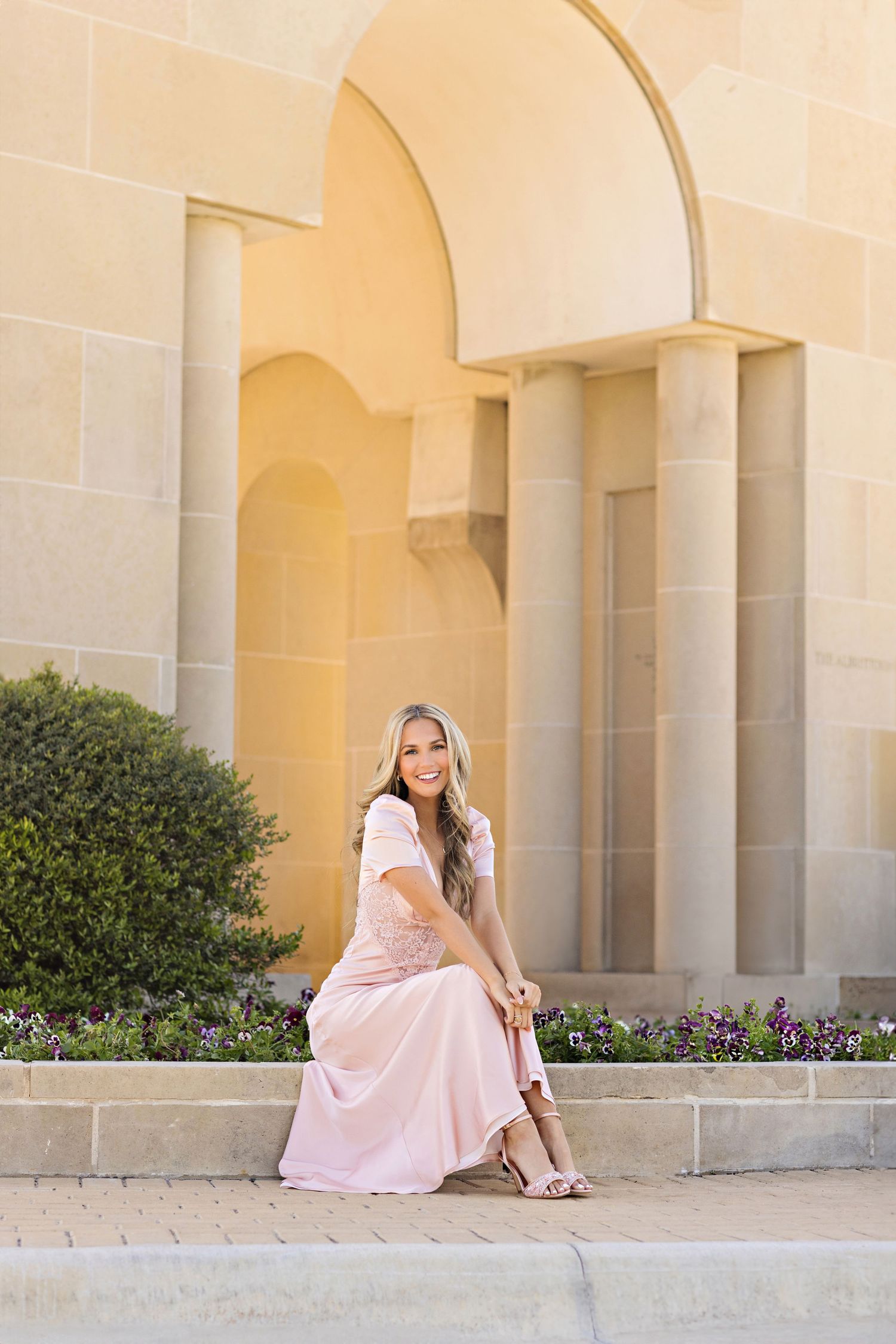 Elegant woman in pink dress sitting on stone steps in front of arched architectural columns at sunset.