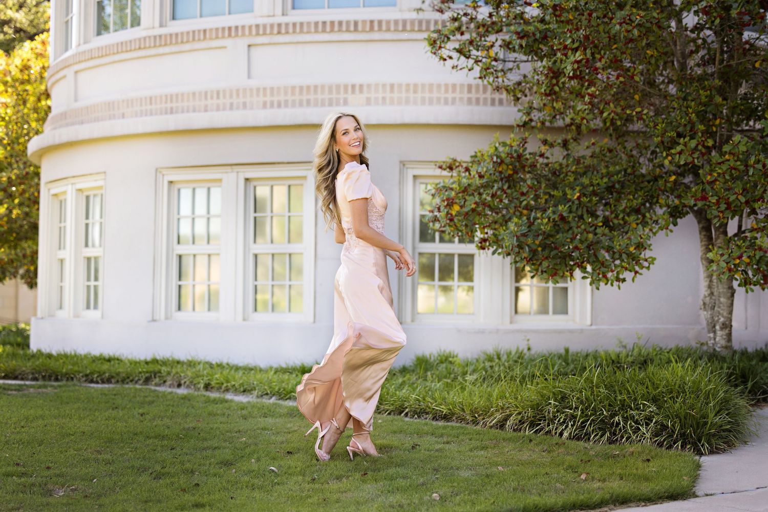 A person in a flowing pink dress twirls gracefully on a lawn in front of an elegant white building.