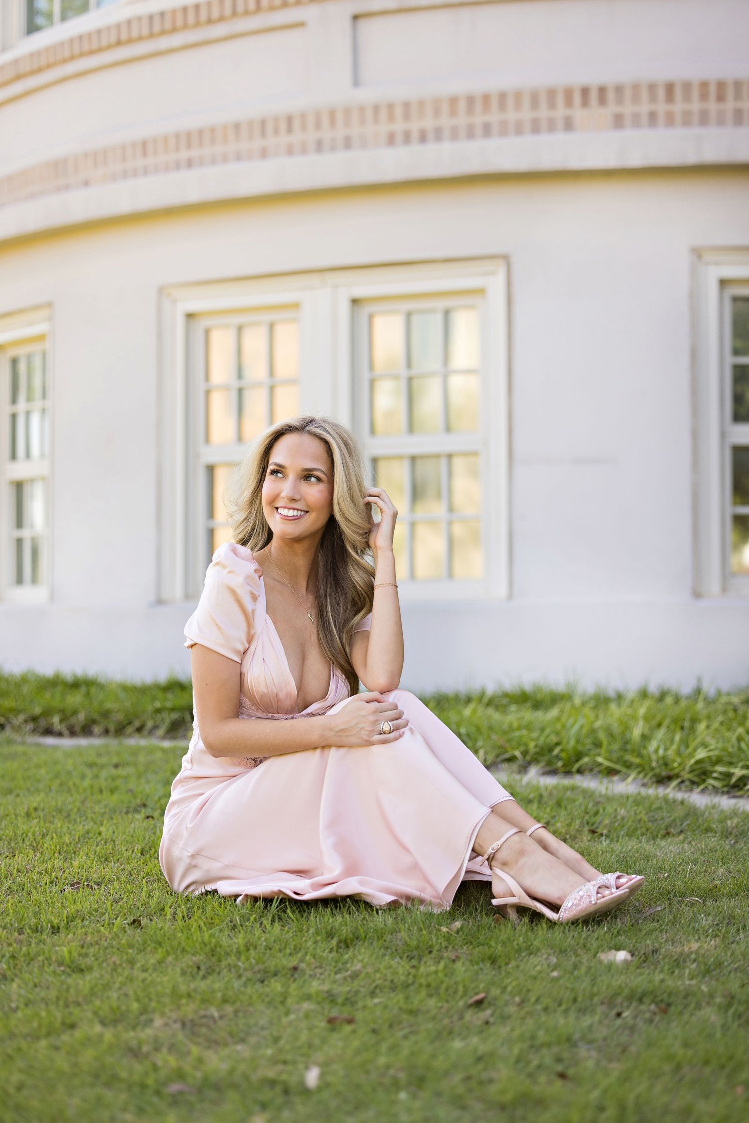 Person in a pink dress sits gracefully on manicured lawn in front of elegant white building with glowing windows.