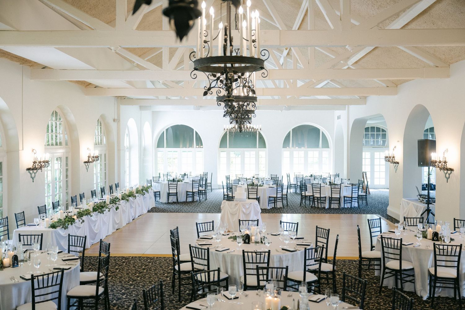 An elegant white reception hall with exposed wooden beams, crystal chandeliers, and Chiavari chairs arranged for an event.