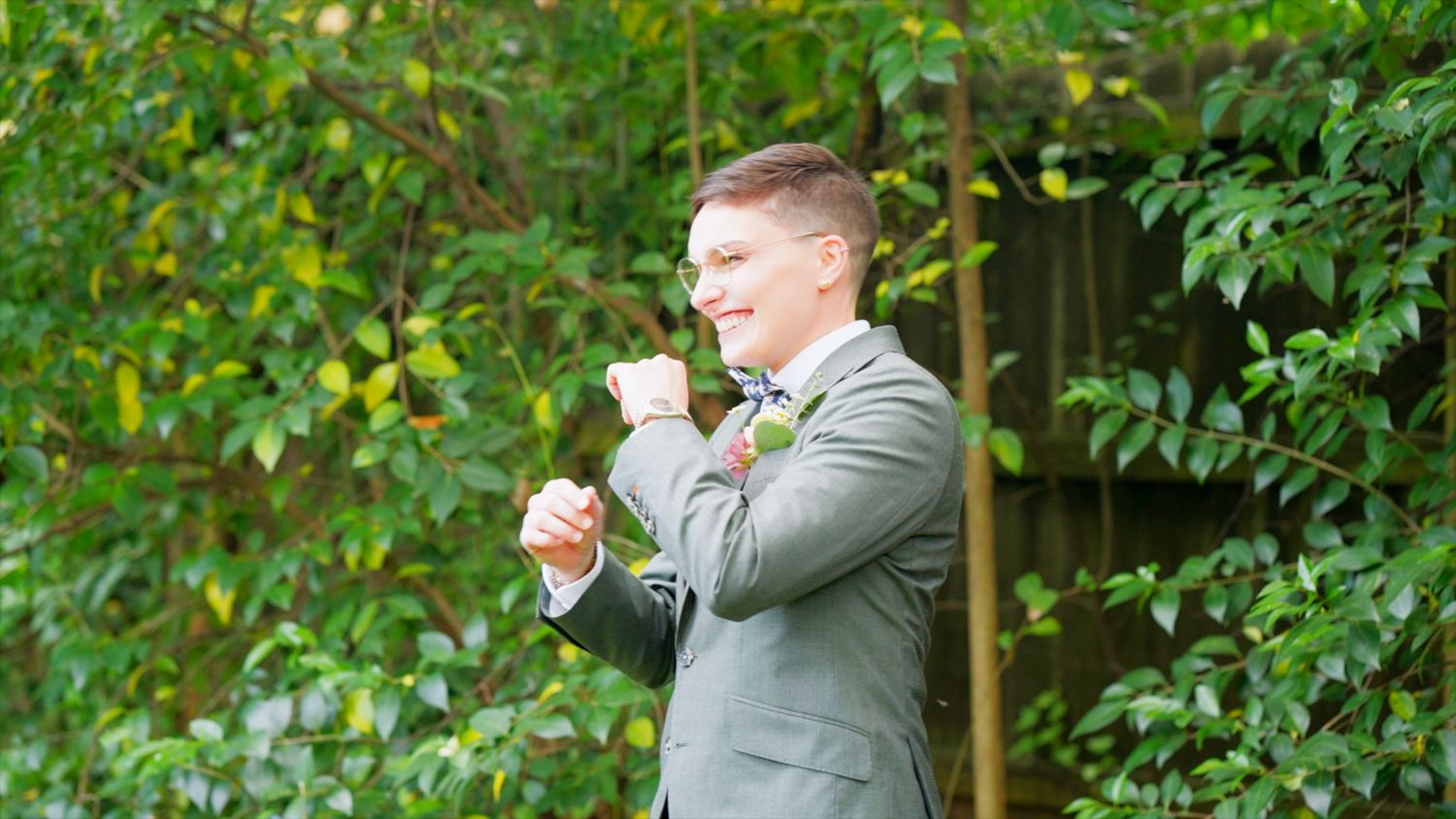 A person in a light grey suit standing outdoors against a backdrop of lush green foliage.