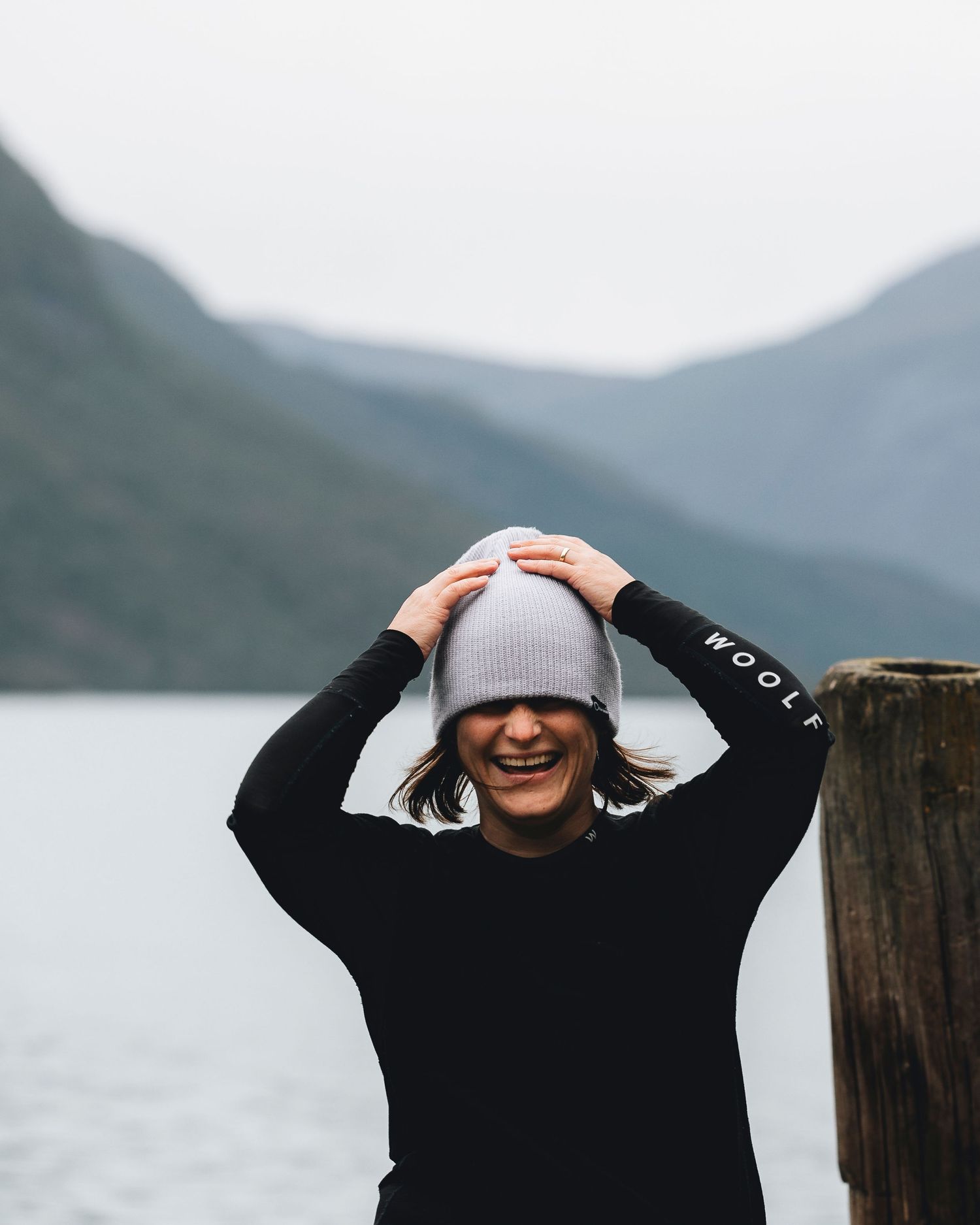 Person wearing a black sweater and gray beanie stands smiling by a lake with mountains in the background.