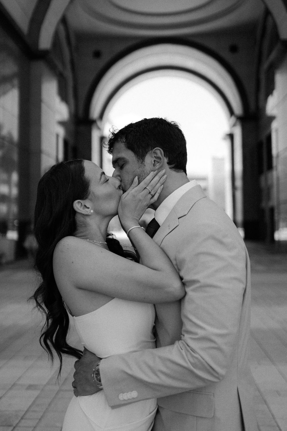 A romantic black and white portrait of a couple sharing a passionate kiss under an ornate arched hallway.