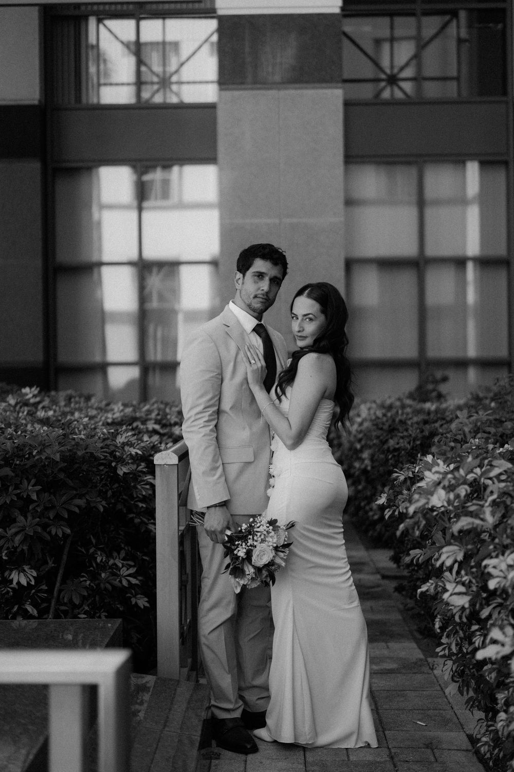 A romantic black and white portrait of newlyweds posing together in formal attire on a garden path with beautiful landscaping.