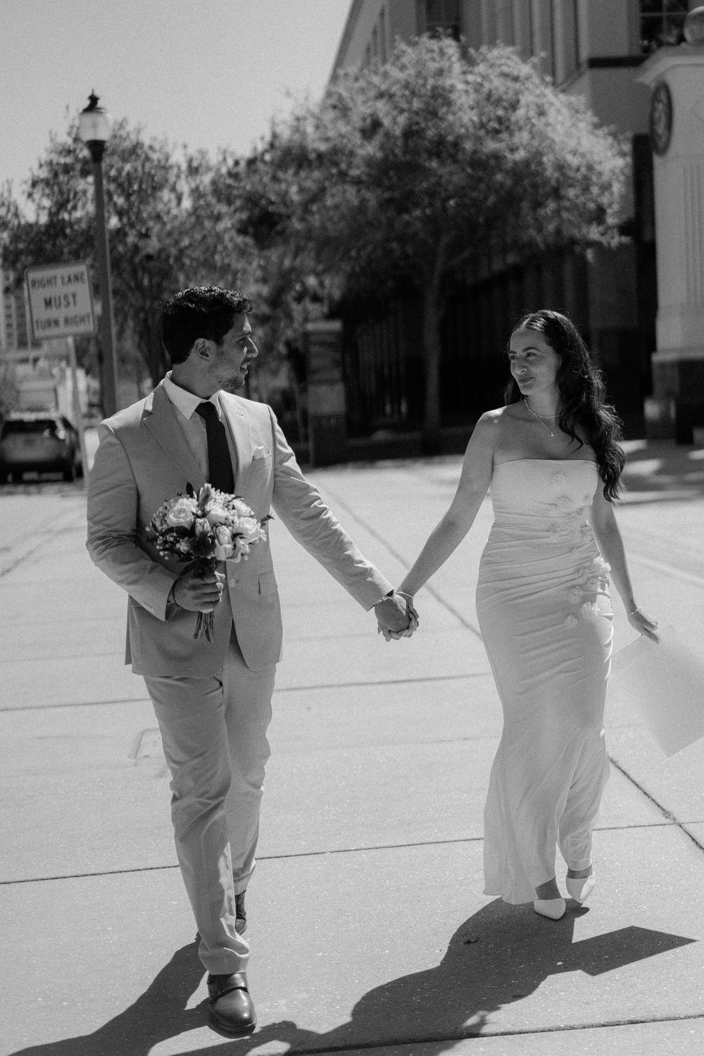Black and white wedding photo of a couple walking down a street together while holding hands and carrying flowers.