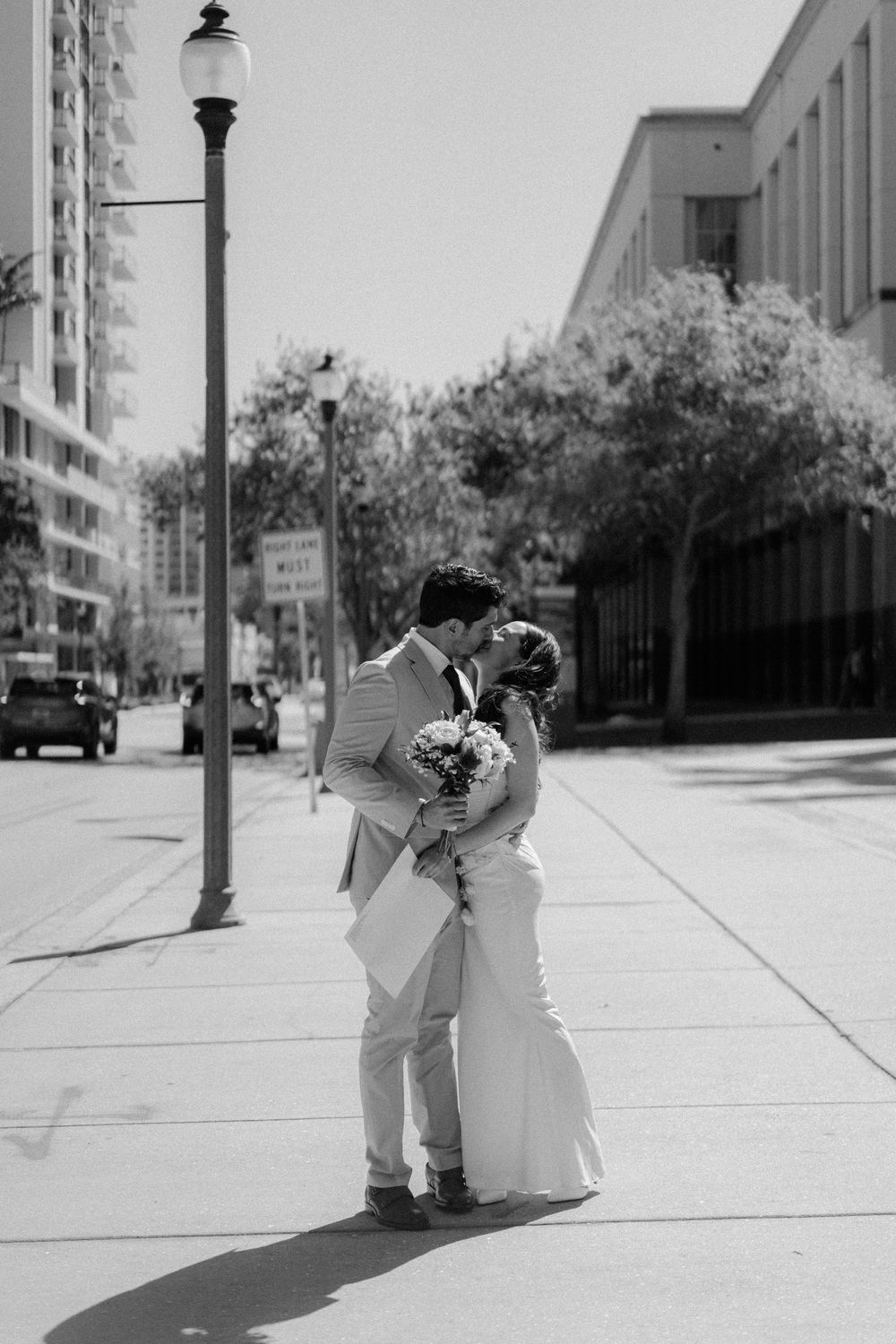 A romantic couple in wedding attire embrace on a sunny urban sidewalk with lamp posts and buildings in view.
