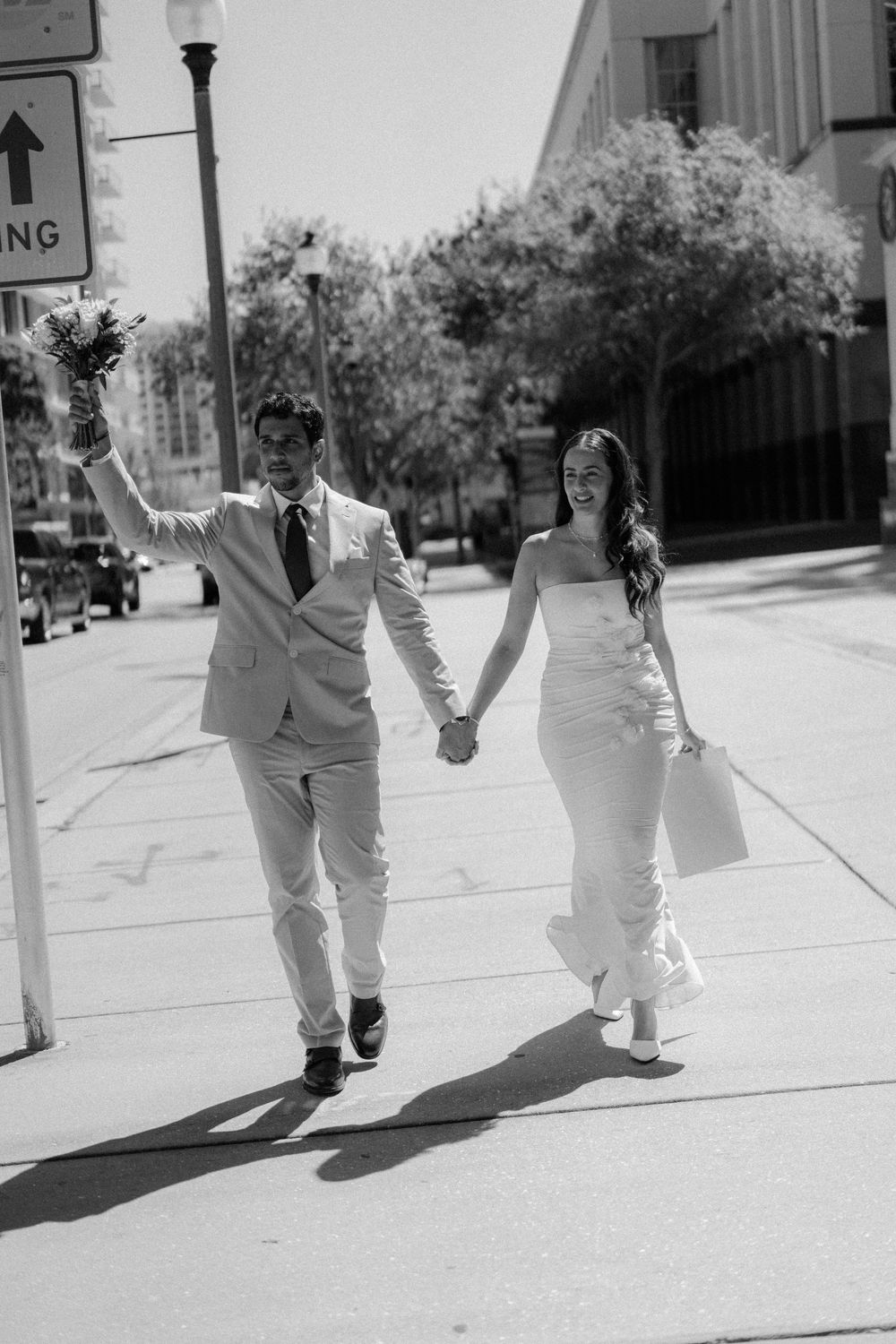 A couple in wedding attire holding hands and walking down a sunny city sidewalk on their special day.