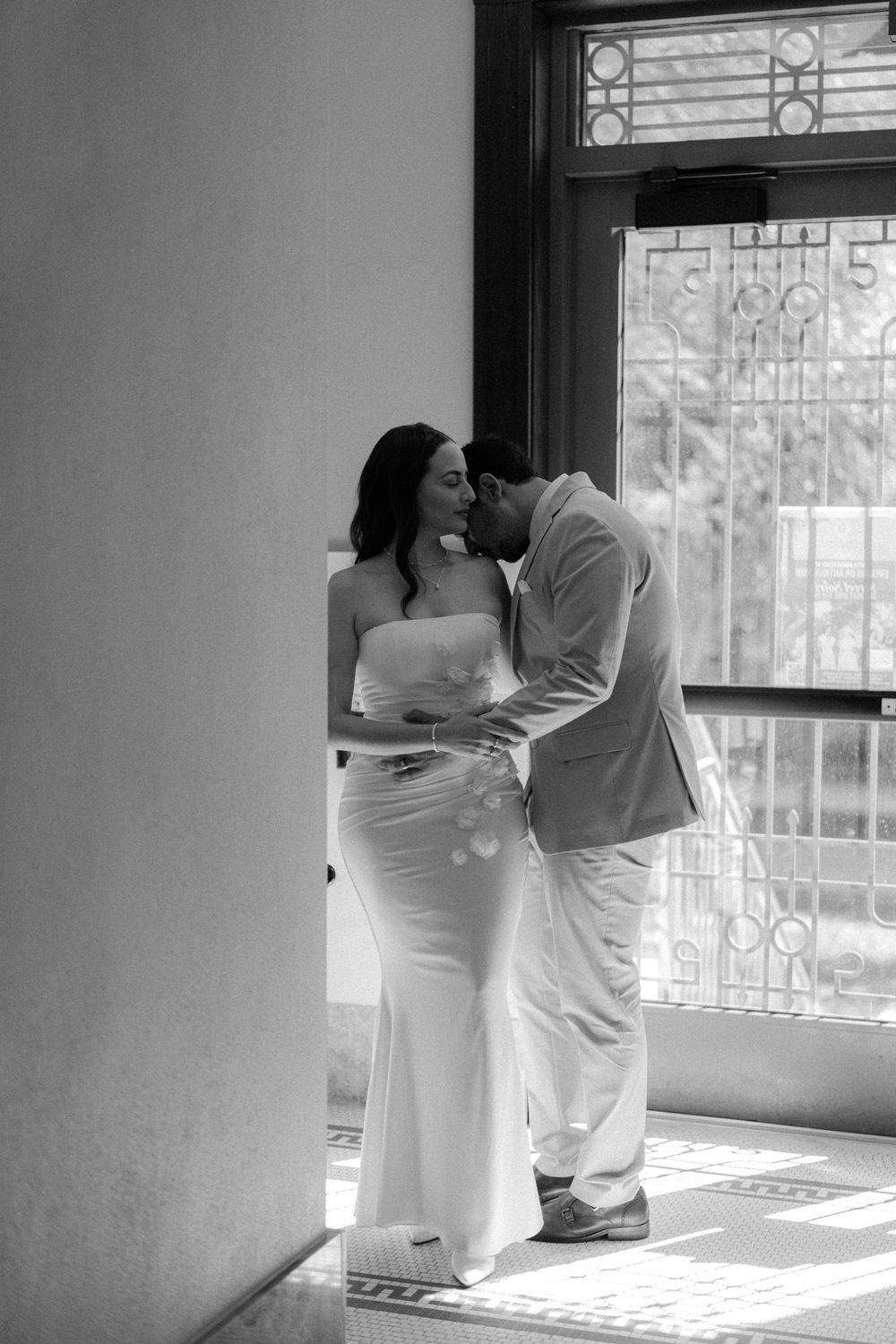 A romantic black and white photo of a couple in elegant attire sharing an intimate moment by a sunlit window.