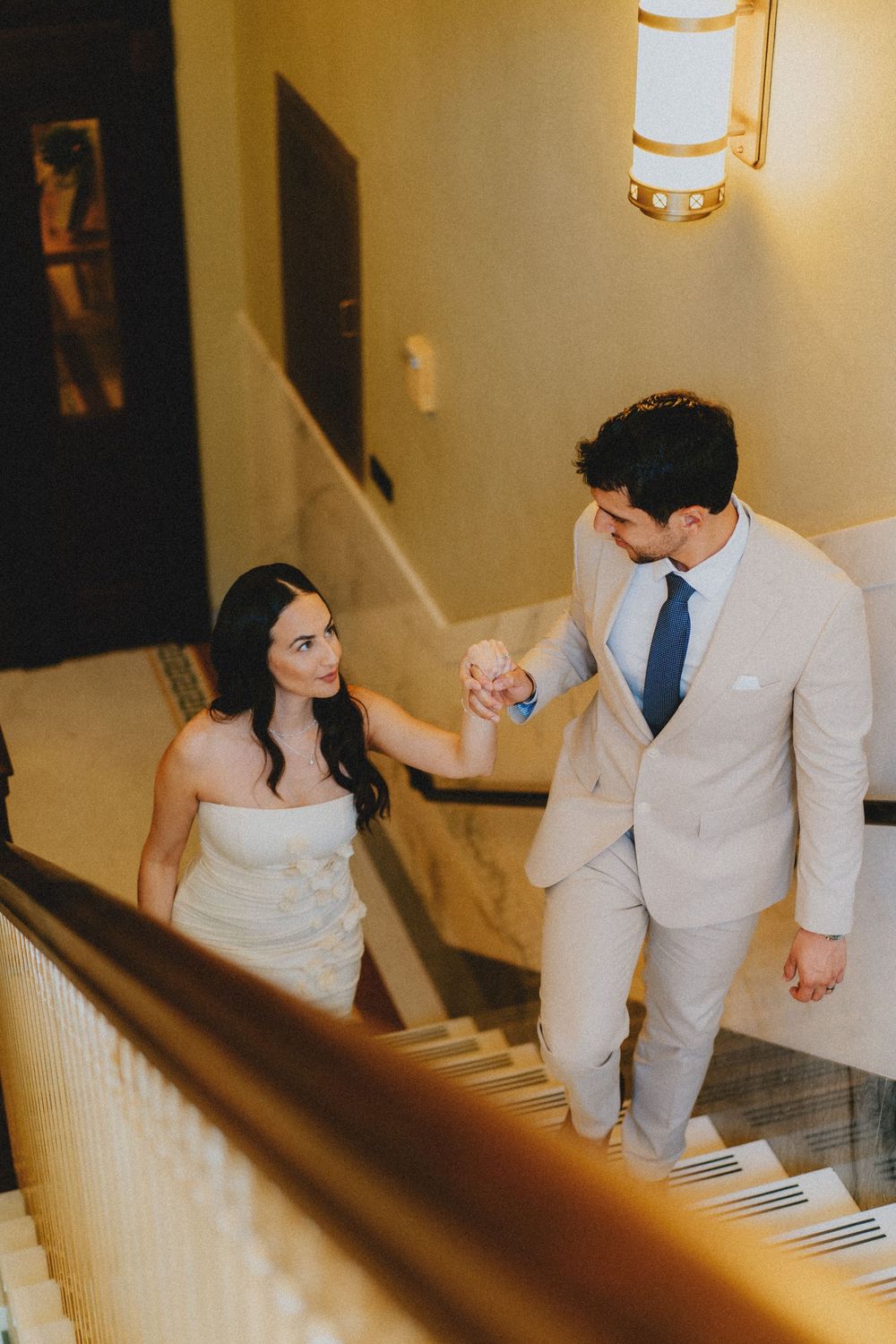 A couple in formal attire walks up an elegant staircase together, with warm lighting illuminating their path.