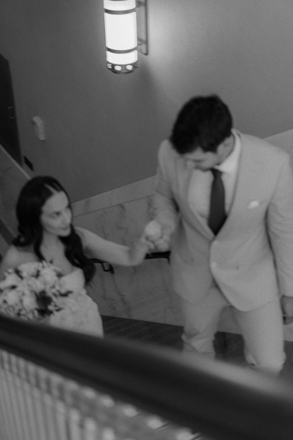 A couple walks up elegant stairs in formal wedding attire as the bride holds a white floral bouquet.