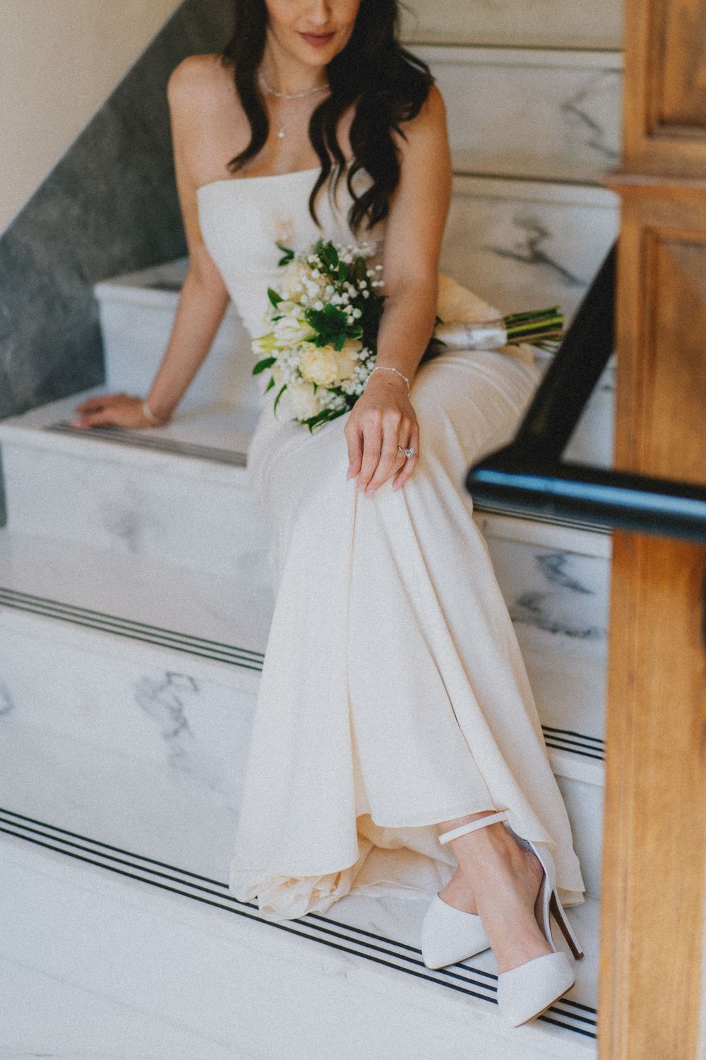 Bride in white wedding dress holding bouquet of lilies while sitting on marble stairs.