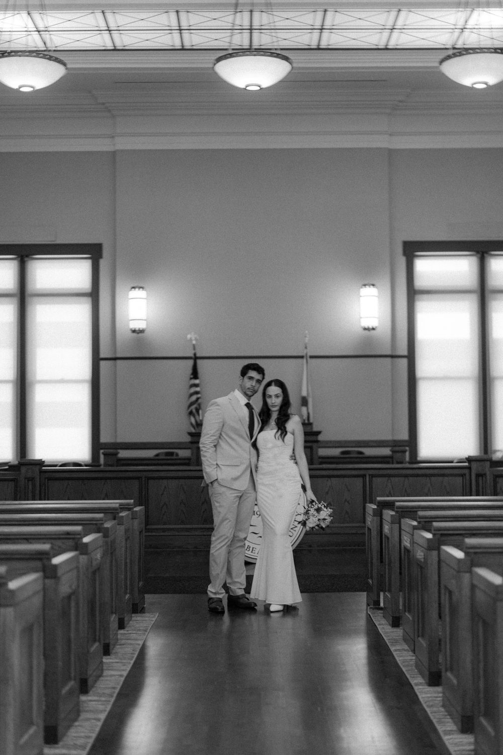 A black and white photo of a wedding couple standing in a traditional church aisle lined with wooden pews.