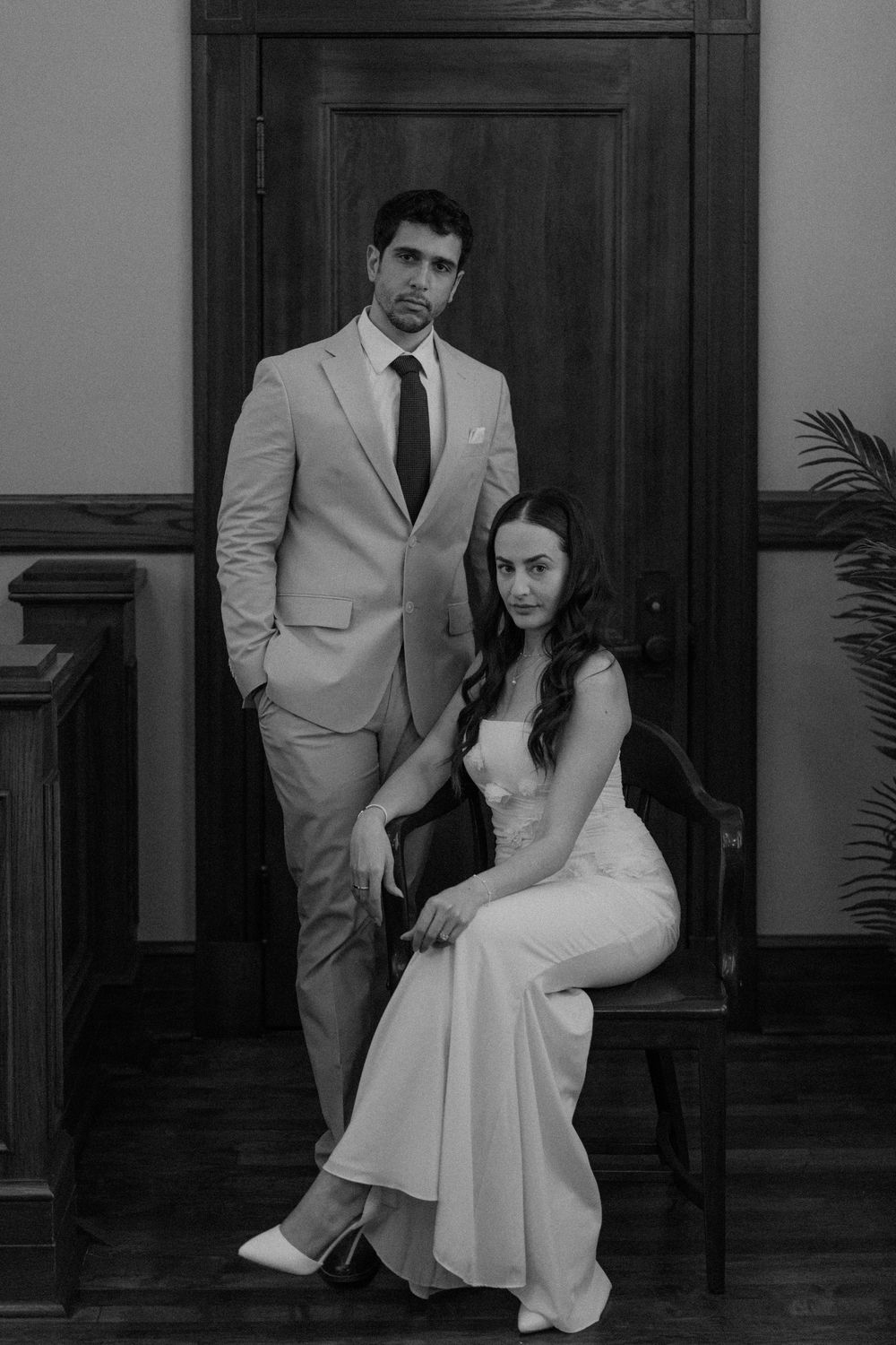 A couple poses formally in black and white, wearing a light suit and white wedding dress while seated on a bench.