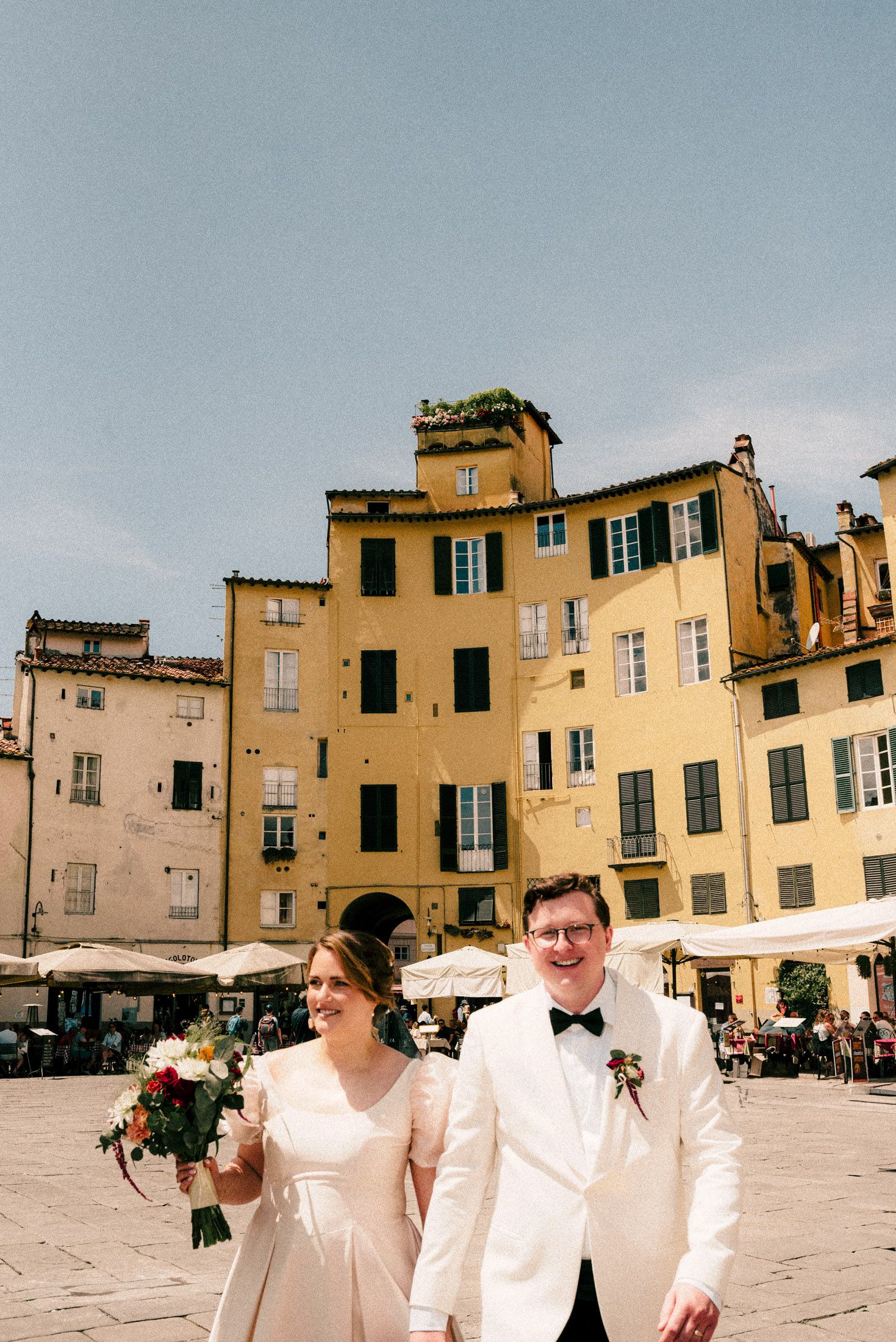 A wedding couple stands in front of historic yellow buildings in an Italian plaza.