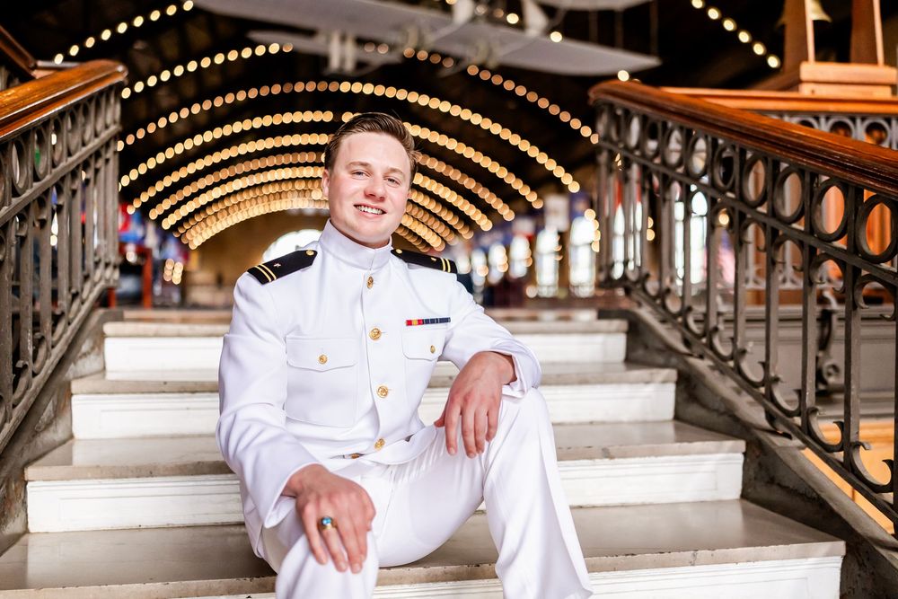 Naval officer in dress whites sits confidently on ornate staircase with illuminated archway above.
