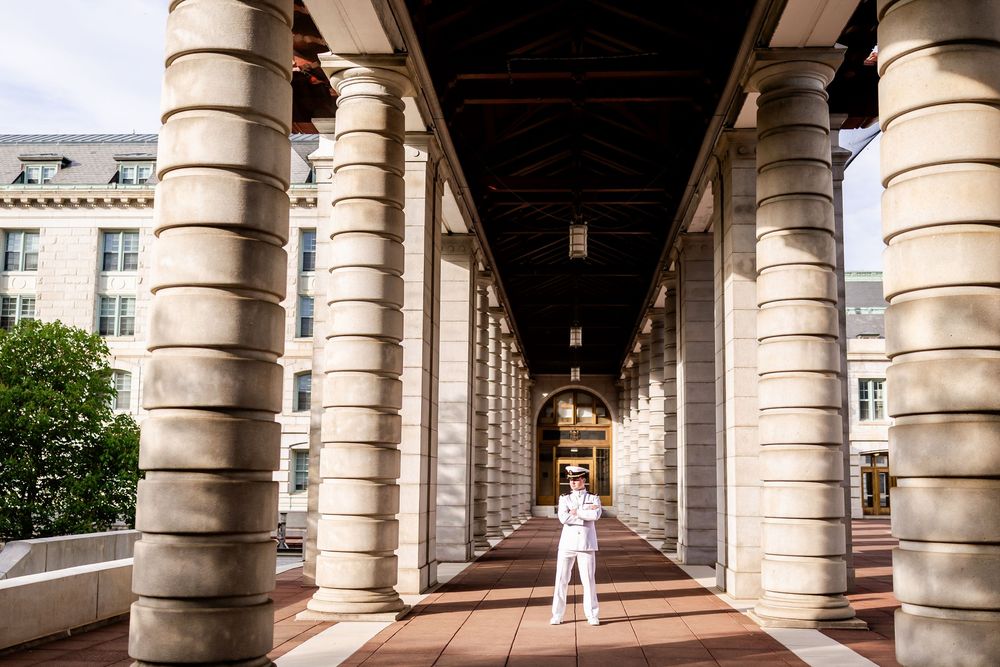 Military officer in dress whites stands under grand architectural columns creating dramatic perspective.