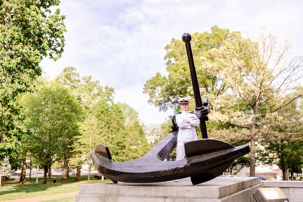 A figure in white naval uniform standing on a large anchor monument in a park setting.