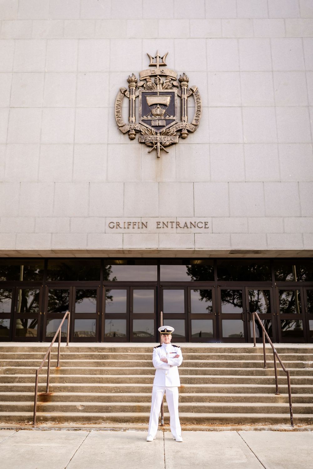 Naval officer in white uniform standing at attention before Griffin Entrance with ornate crest above.