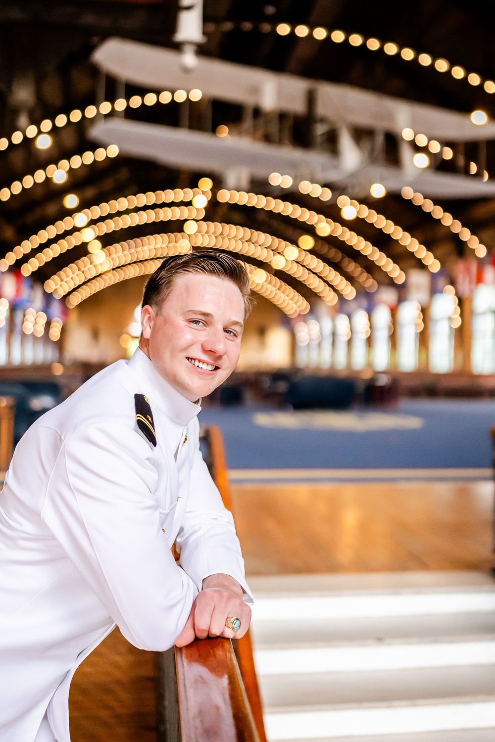 Naval officer in white uniform smiling under decorative string lights in an elegant venue.