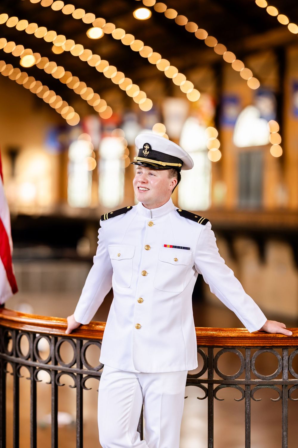 Naval officer in dress whites poses against backdrop of decorative lights and American flag.
