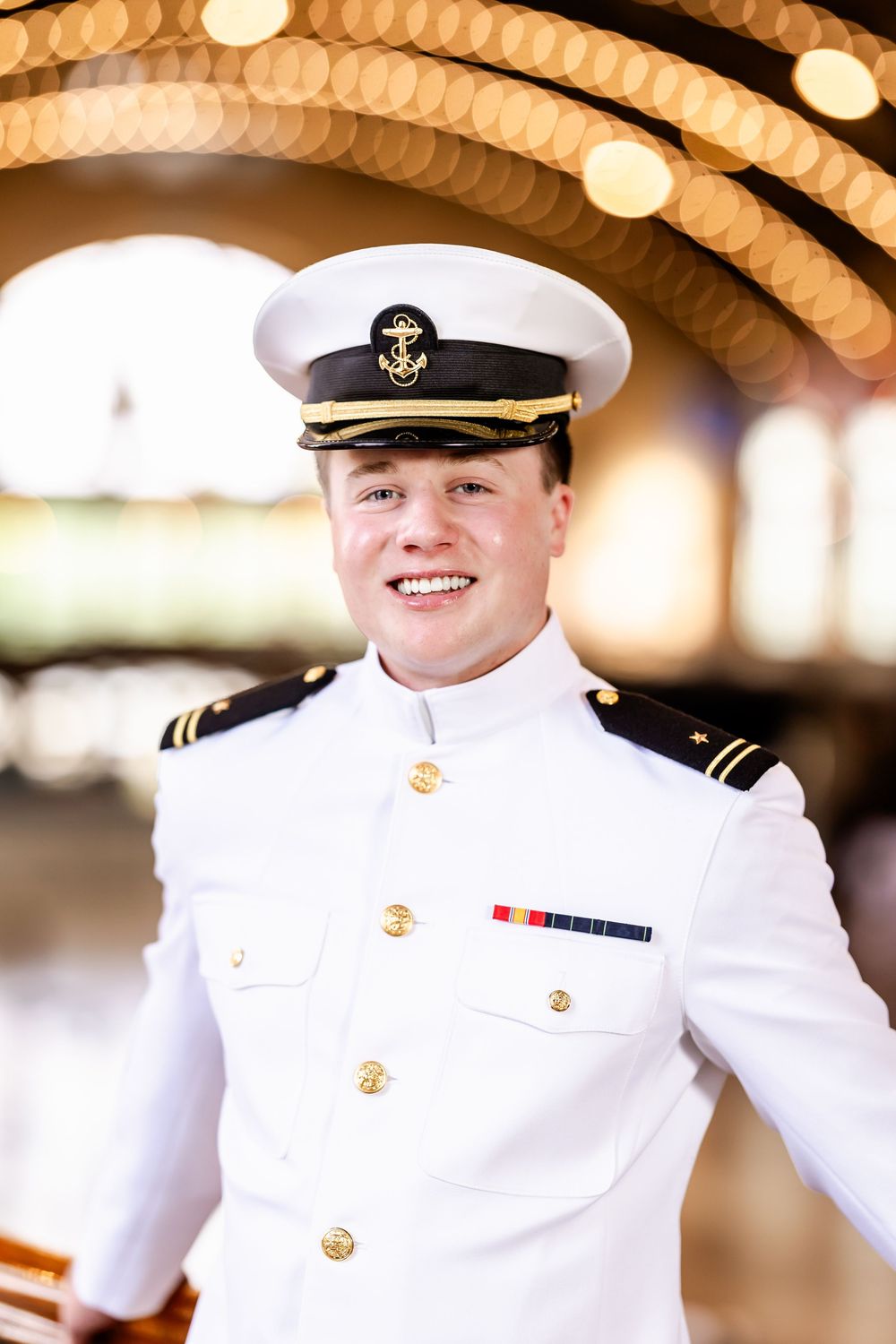 Naval officer in dress whites with cap smiling beneath string lights.