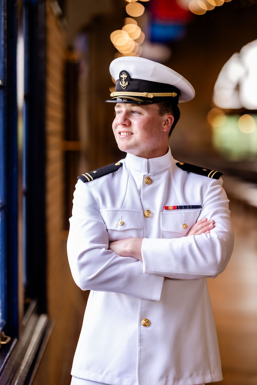 Naval officer in white uniform with arms crossed standing indoors.