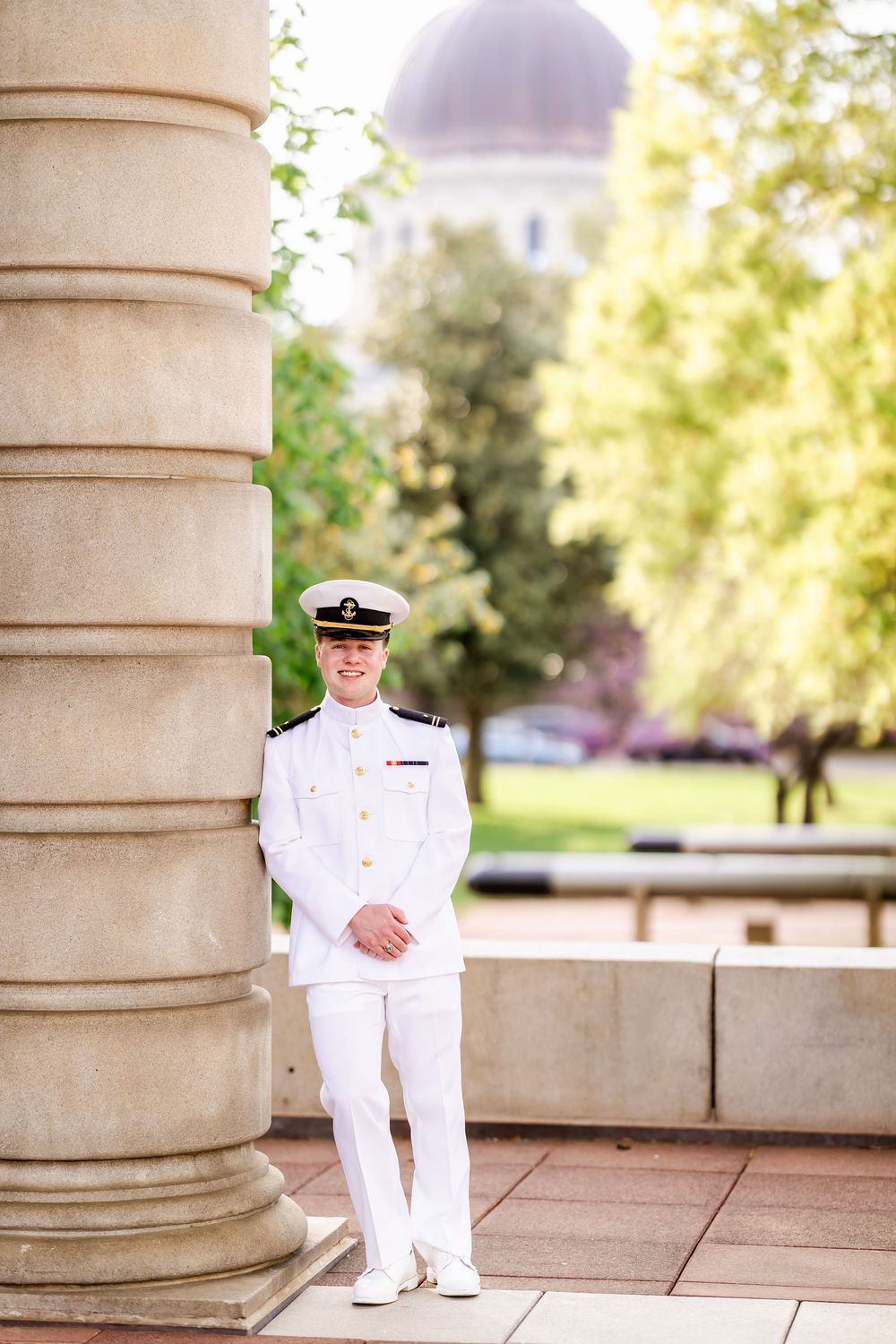 Naval officer in dress whites standing beside classical column with dome visible in background.