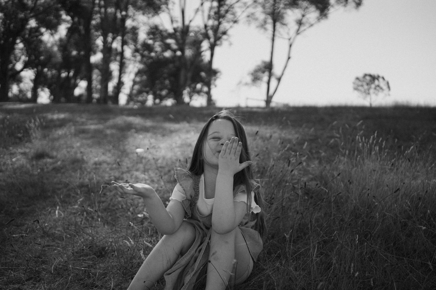 Black and white portrait of person sitting in grassy field blowing bubbles with trees in background.