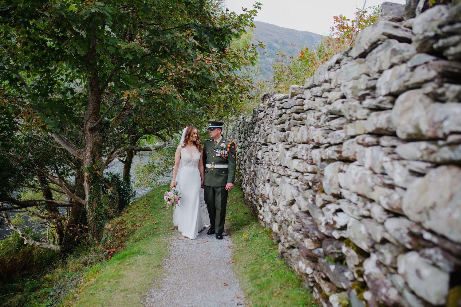 Two people walk together on a scenic mountain path beside a rustic stone wall in a green landscape.