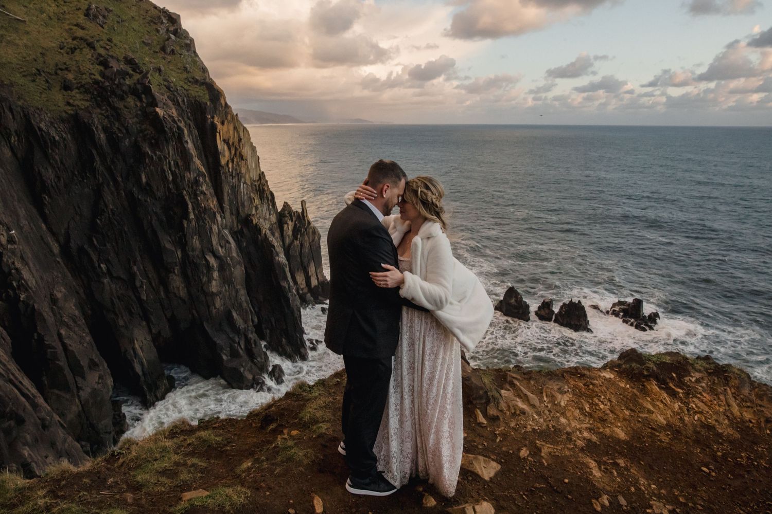 A couple embraces on a dramatic cliff edge overlooking the ocean at sunset, with rugged coastal rocks in the background.