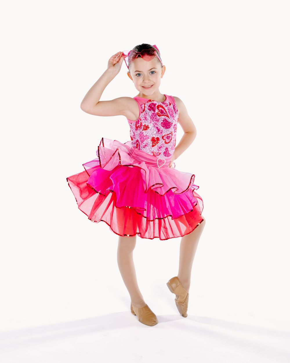 Young dancer in pink floral costume with ruffled tutu performs graceful dance move against white background.