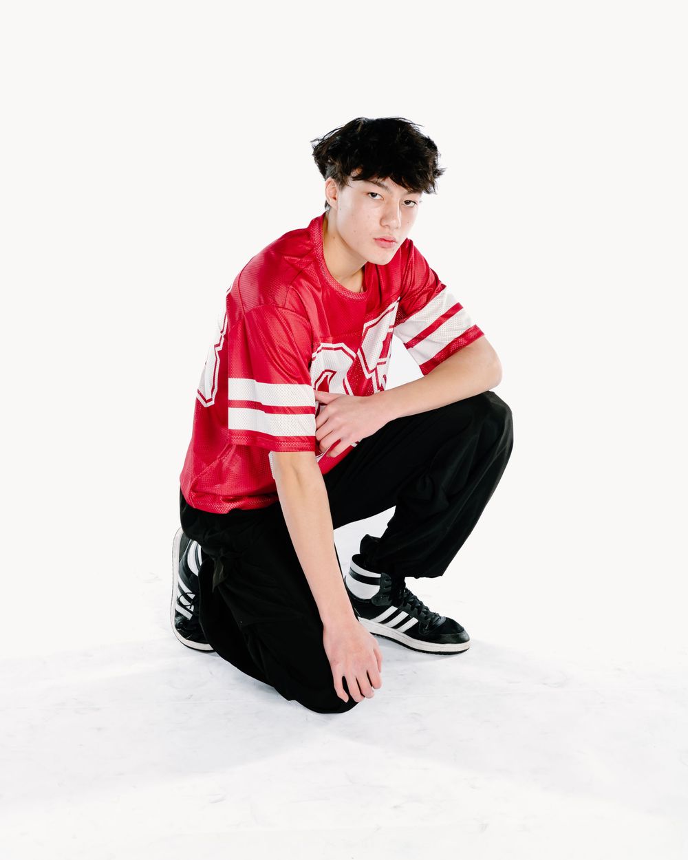 A person in a red and white sports jersey and black pants crouches while wearing athletic sneakers.