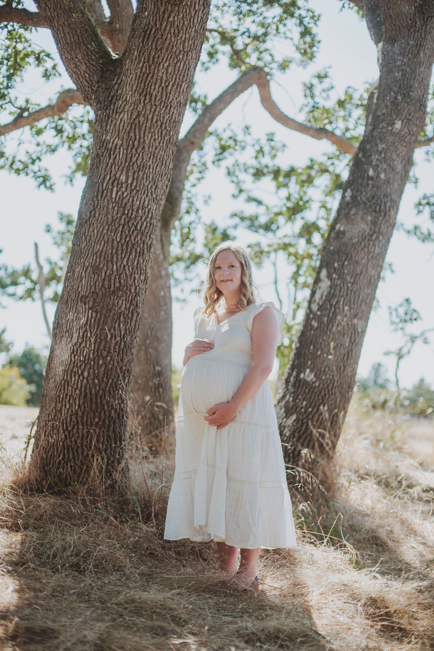 A pregnant woman in a flowing white maternity dress stands between large oak trees in soft sunlight.
