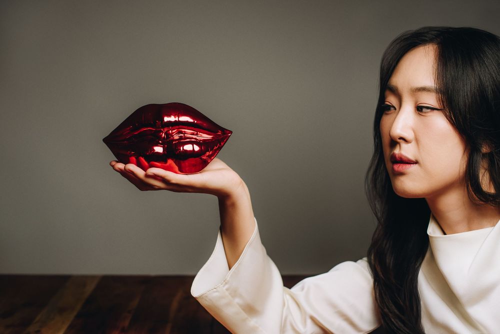 Person in white sweater holds shiny red decorative lips sculpture against neutral background.