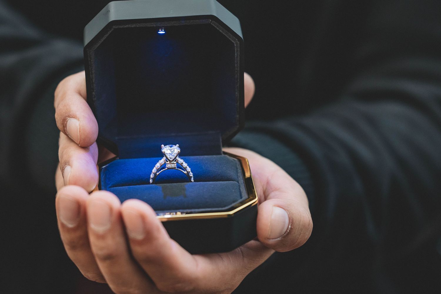 A diamond engagement ring sits in a blue velvet ring box held by hands.
