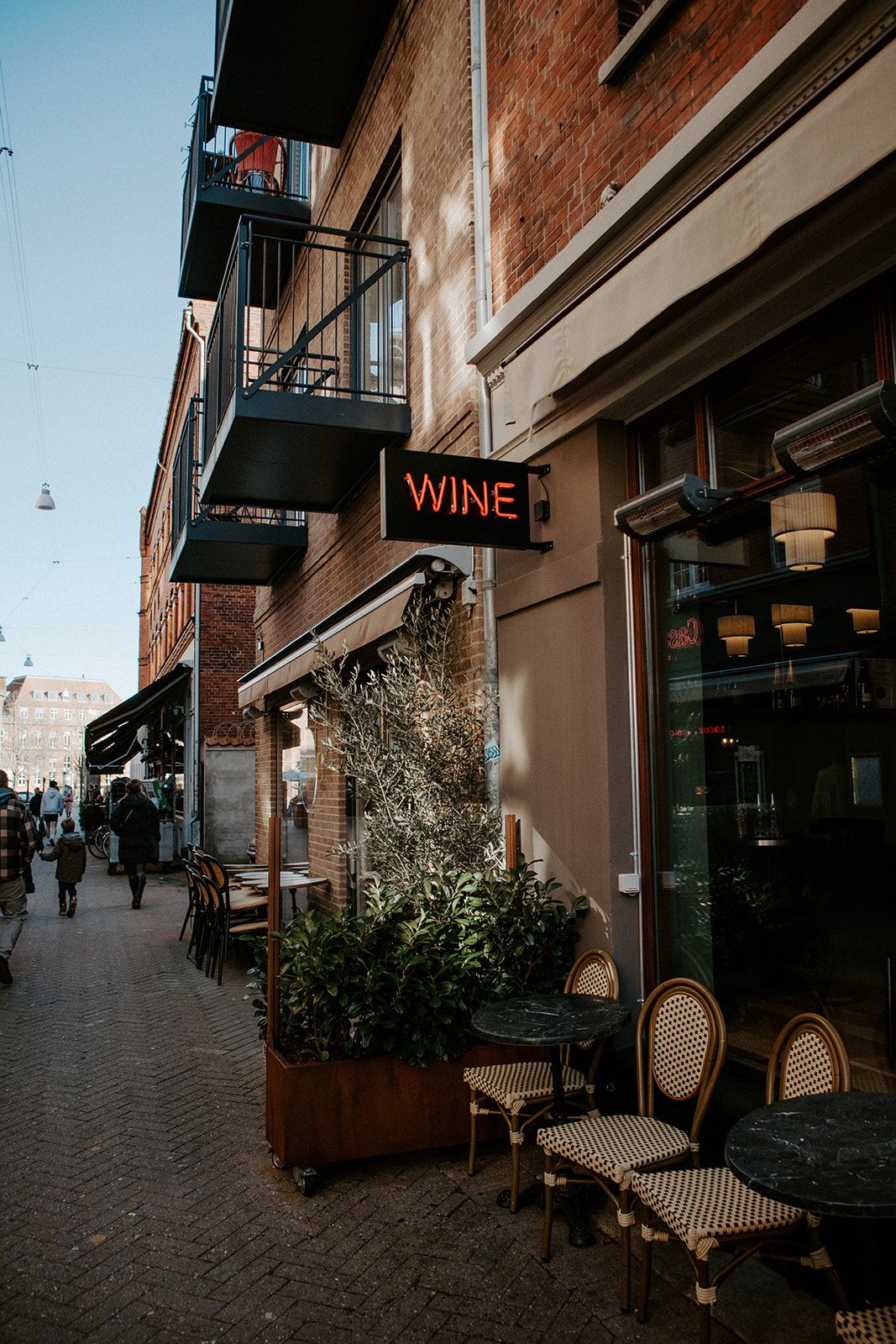 street photography på en gade i Odense by på Fyn, foran en fransk-inspireret vin butik med rødt neon skilt og fletstole på gaden