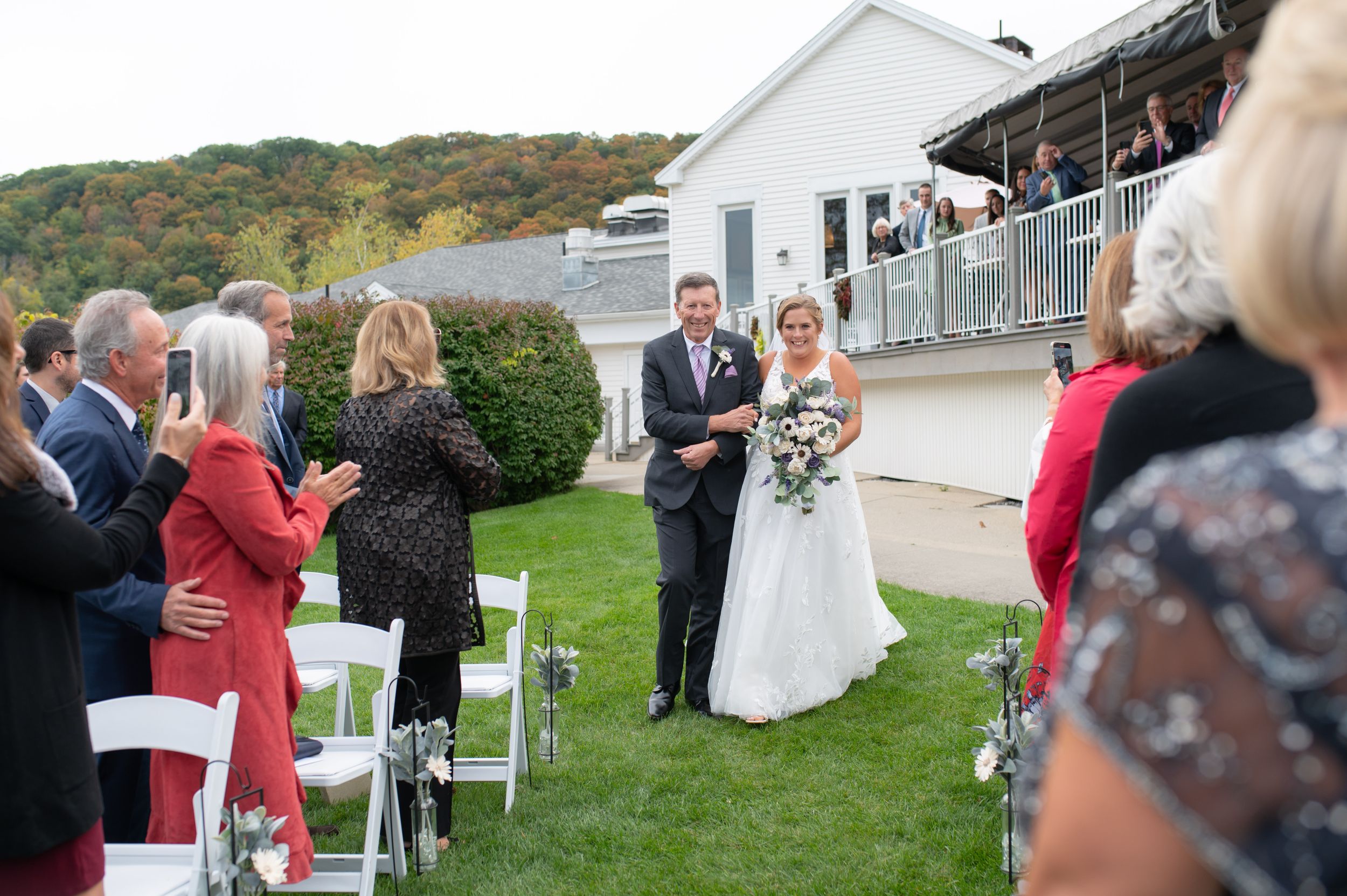 Wedding ceremony guests gather outdoors on a lawn with white chairs and scenic hillside backdrop.