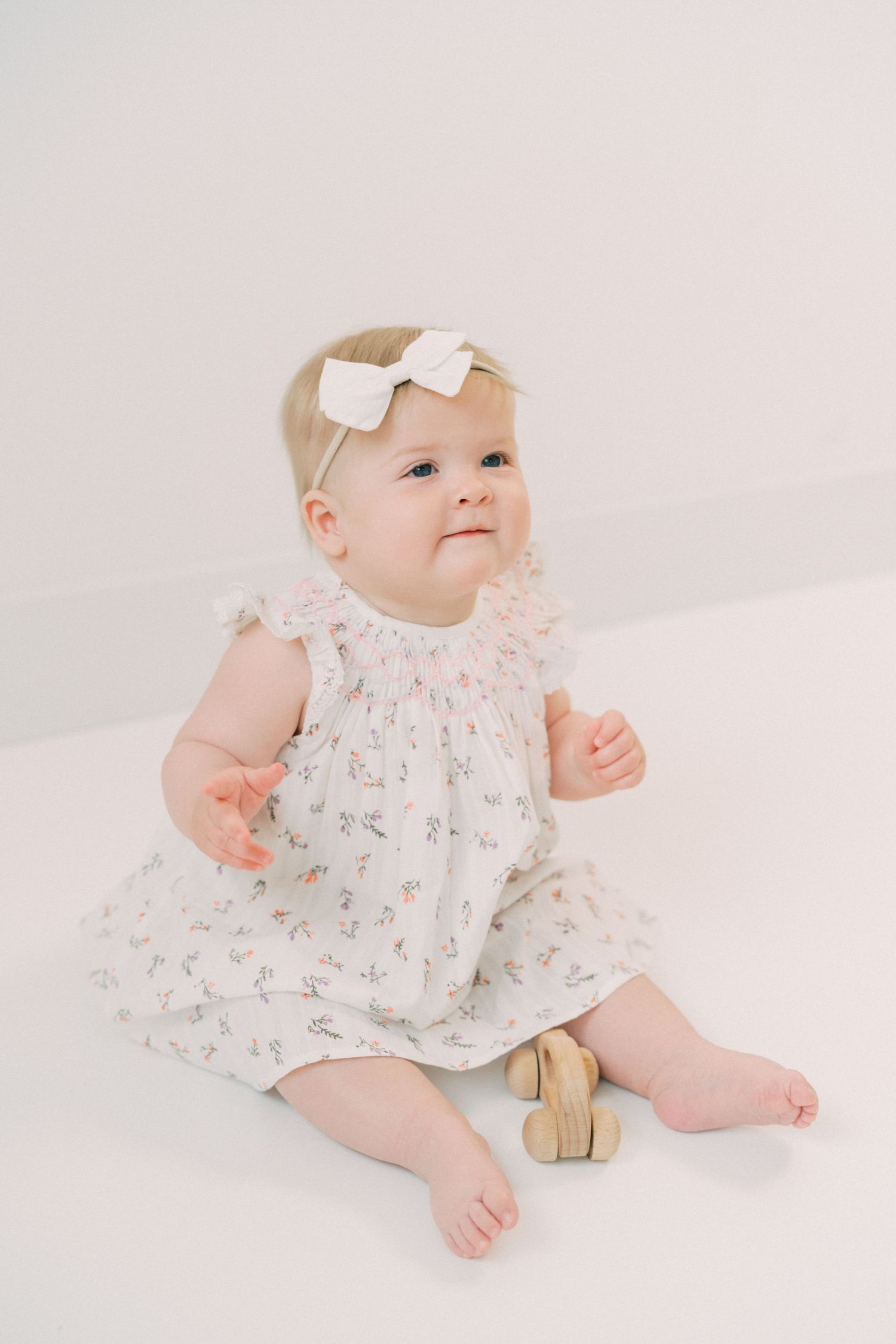 Baby in floral dress with white bow sitting on white backdrop wearing tan shoes and gazing upward.