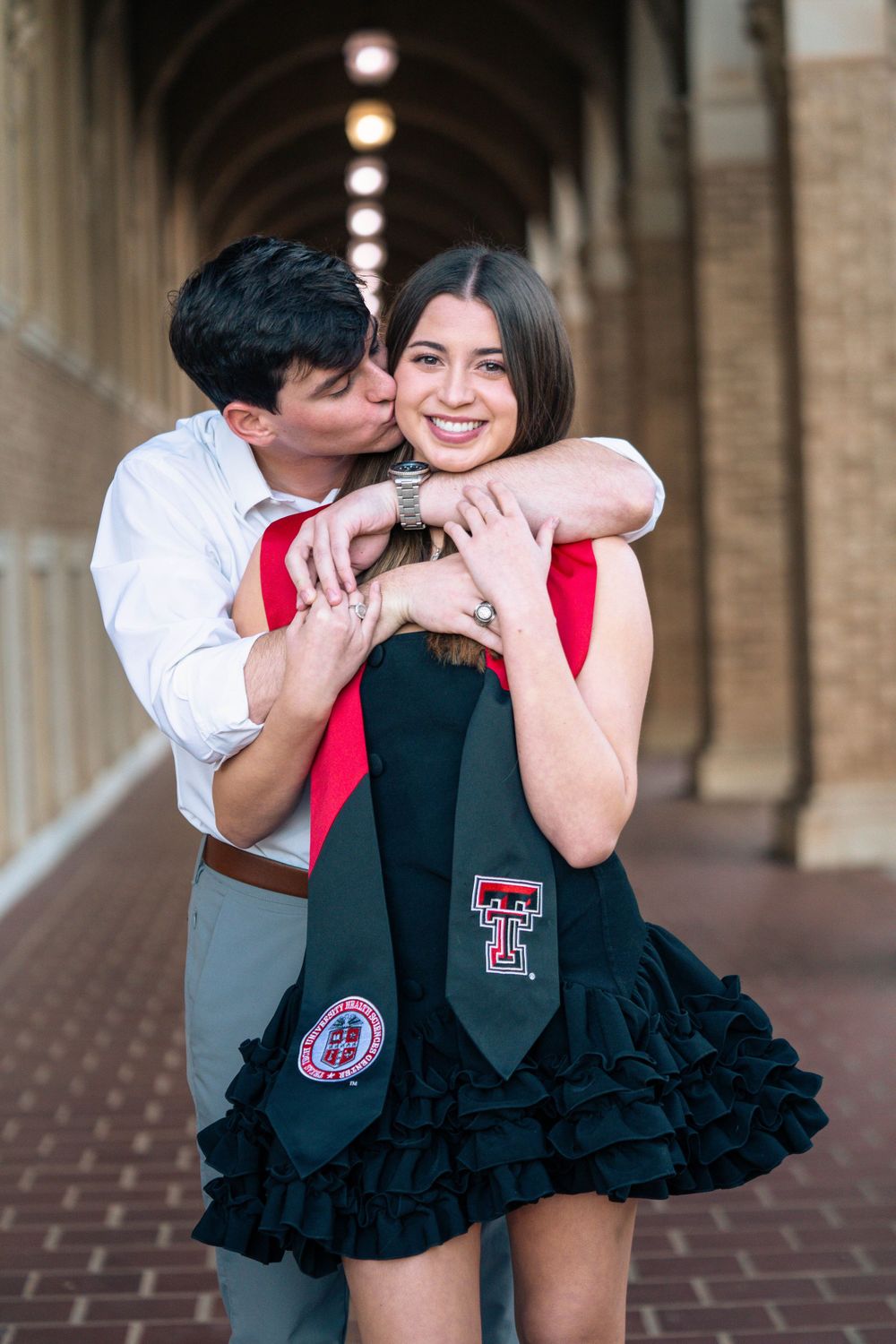 Two people in graduation attire share a candid moment in front of arched brick columns.