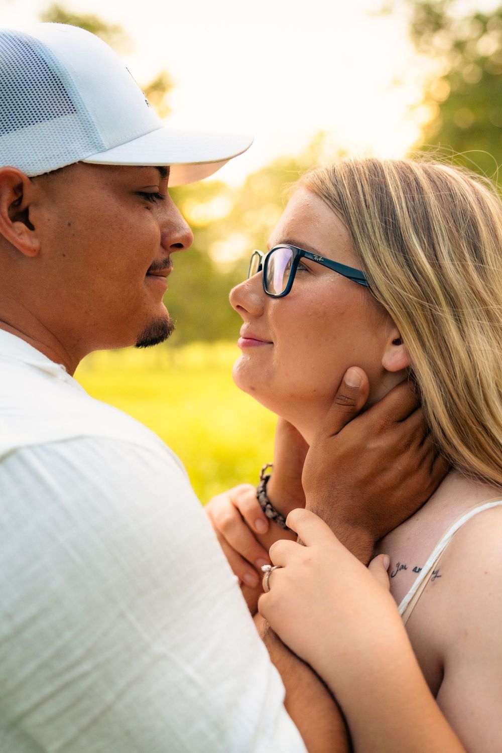 Romantic couple shares intimate moment in golden sunlight as she wears glasses and he wears a white baseball cap.