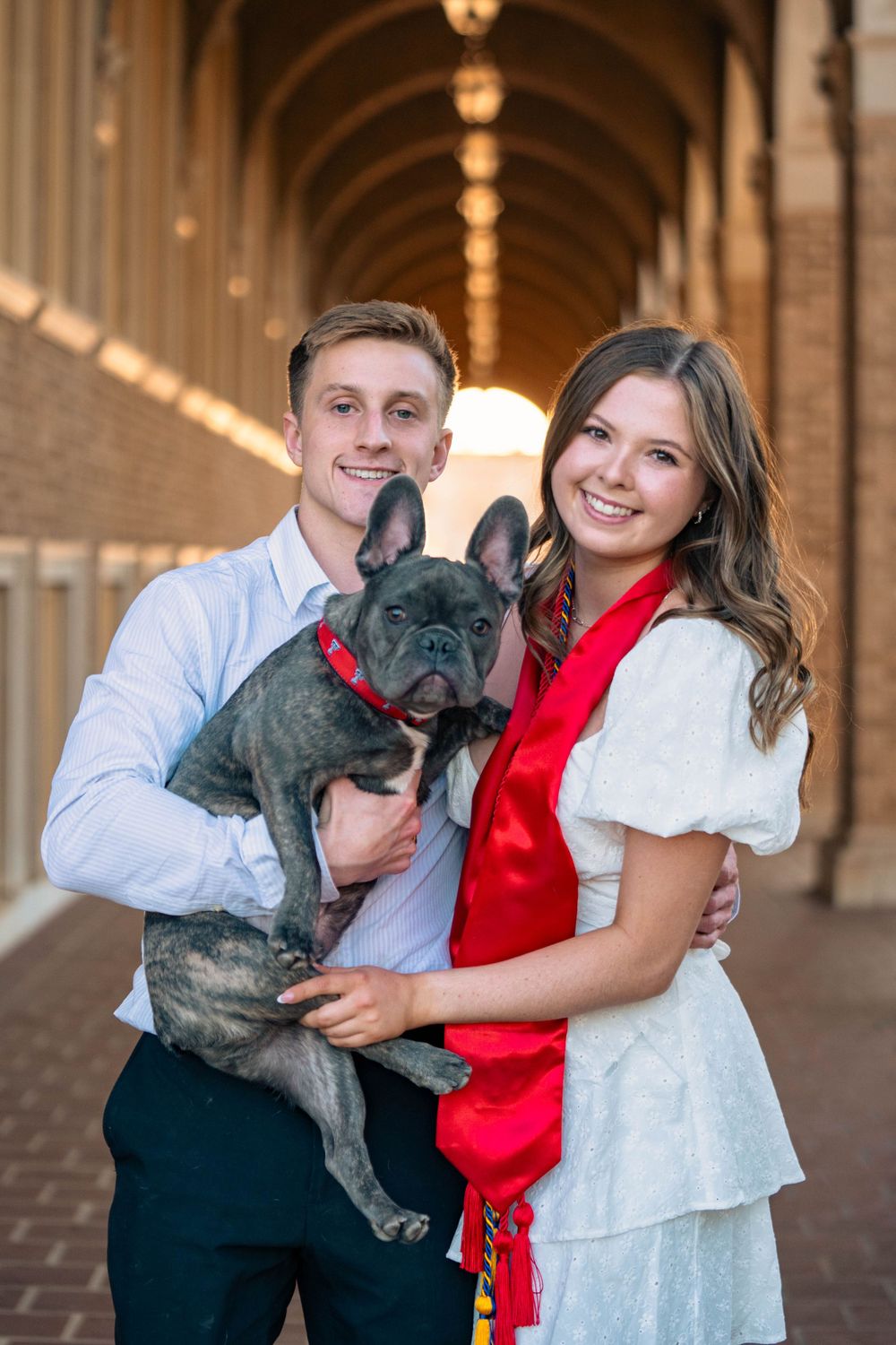 Couple poses with their French Bulldog in formal attire under an ornate brick colonnade.