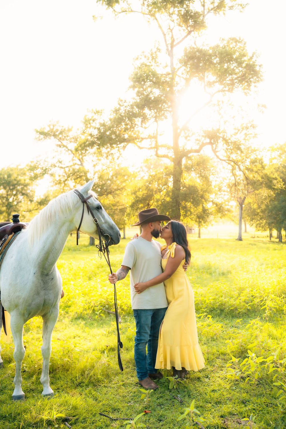 A romantic photoshoot in a sunlit meadow features a couple in yellow and white posing with a majestic white horse at sunset.