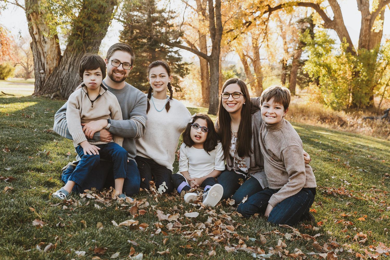 A happy family sits together on autumn grass surrounded by fallen leaves during a golden sunset in the park.