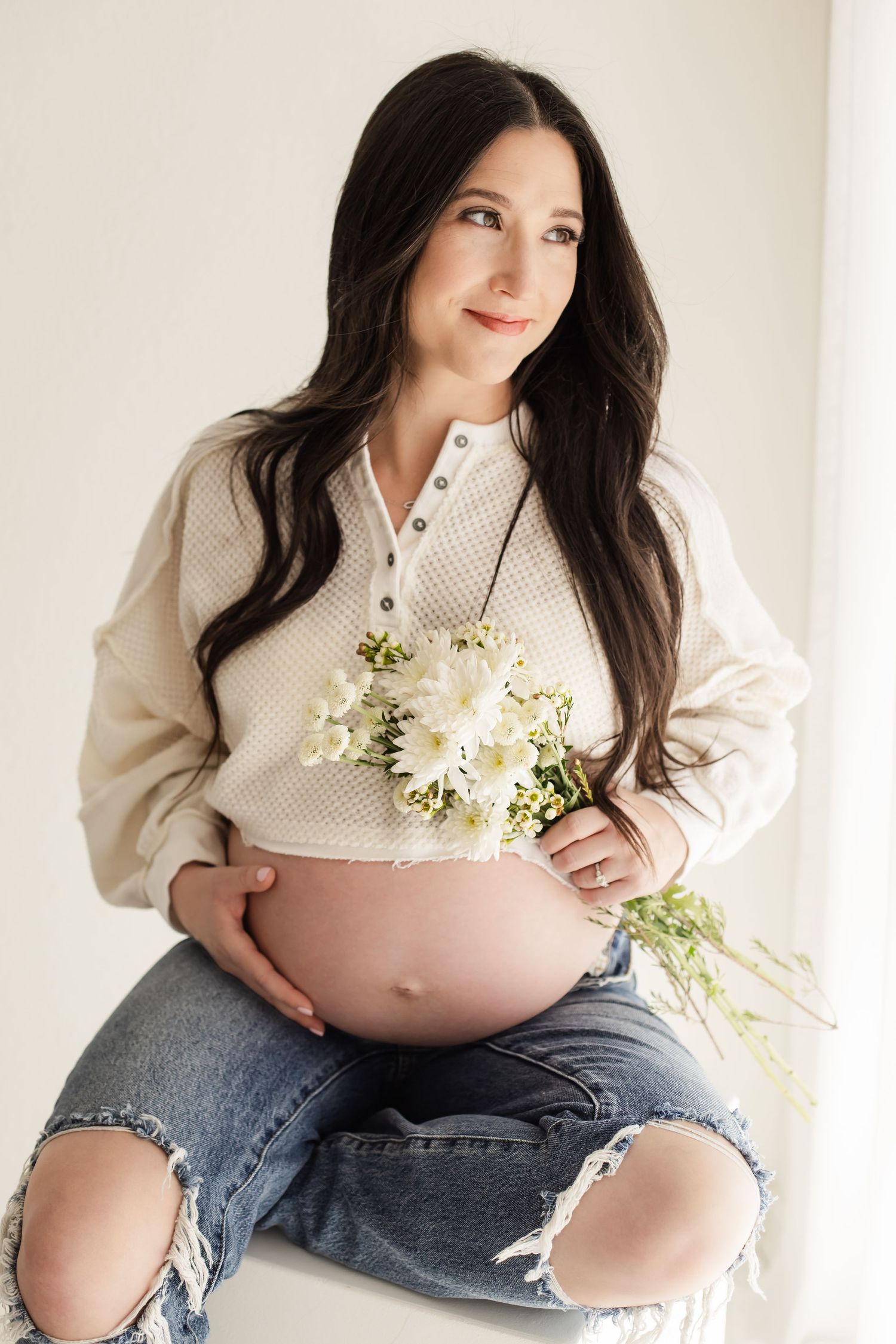 Simple and comfortable maternity portrait of a pregnant mom staring out the window holding florals in the white studio room.