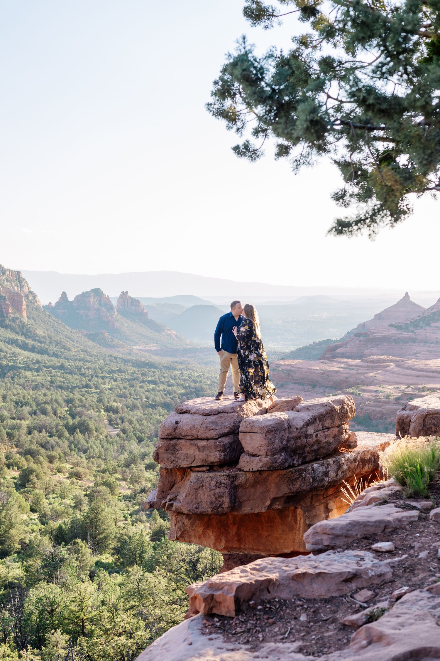 A couple and their dog stand on a rocky outcrop overlooking Sedona's dramatic red rock formations.